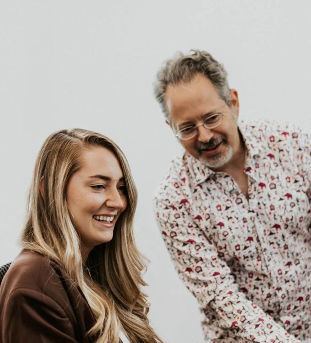 A woman seated at a desk with a smiling man standing beside her. Both are looking at something off-screen.
