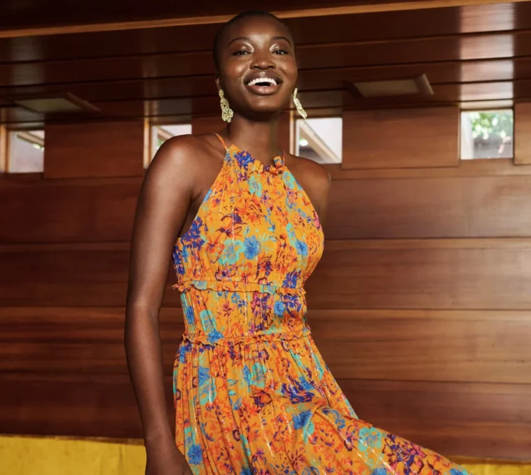 Person in an orange floral dress stands inside a wood-paneled room, smiling with one foot slightly raised.