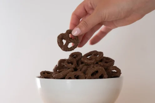 A hand holding a chocolate-covered pretzel above a white bowl filled with similar pretzels, against a plain background.