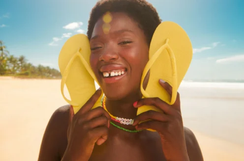 Person smiling on a beach, holding yellow flip-flops to their cheeks. Wearing a colorful swimsuit and necklace, with palm trees and blue sky in the background.