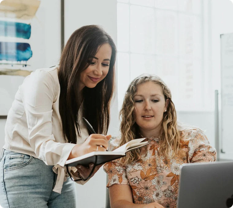 Two women working together; one is writing in a notebook, the other is looking at a laptop screen in an office setting.