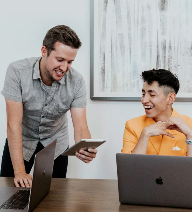 Two people at a table with laptops, one standing and holding a tablet, the other seated and smiling. A framed abstract artwork hangs on the wall behind them.