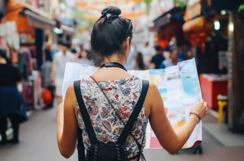 A woman stands in a bustling market street, holding a map. She has a backpack and is wearing a floral top, with her hair in a bun. The market around her is busy and colorful.