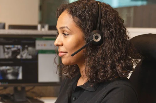 A woman with curly hair is wearing a headset and sitting at a desk with computer monitors.