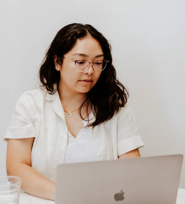 Person with glasses sitting at a table, looking at a laptop screen.