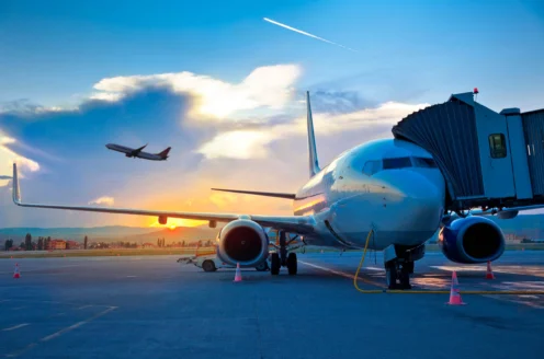 Airplane parked at the airport gate with a jet bridge attached, another plane taking off in the background against a sunset sky.