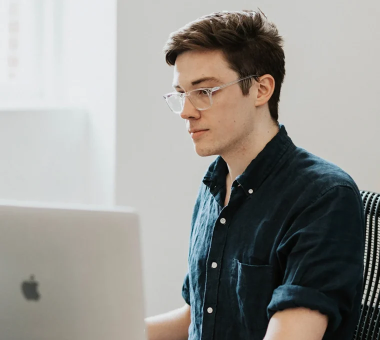 Person with glasses working on a laptop at a desk, wearing a dark shirt, in a well-lit room.