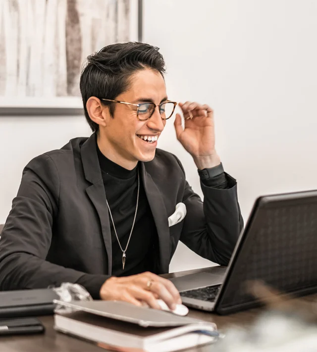 A person in a black blazer and glasses smiles while working on a laptop at a desk with books and papers.