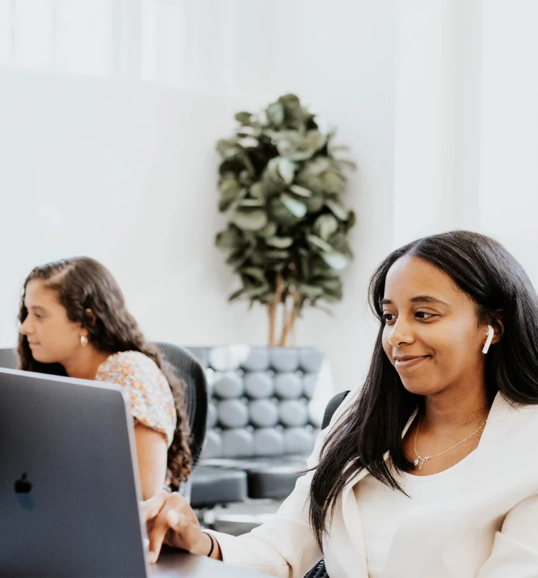 Two people work at laptops in a bright office with a large plant in the background. One person wears earphones and smiles.