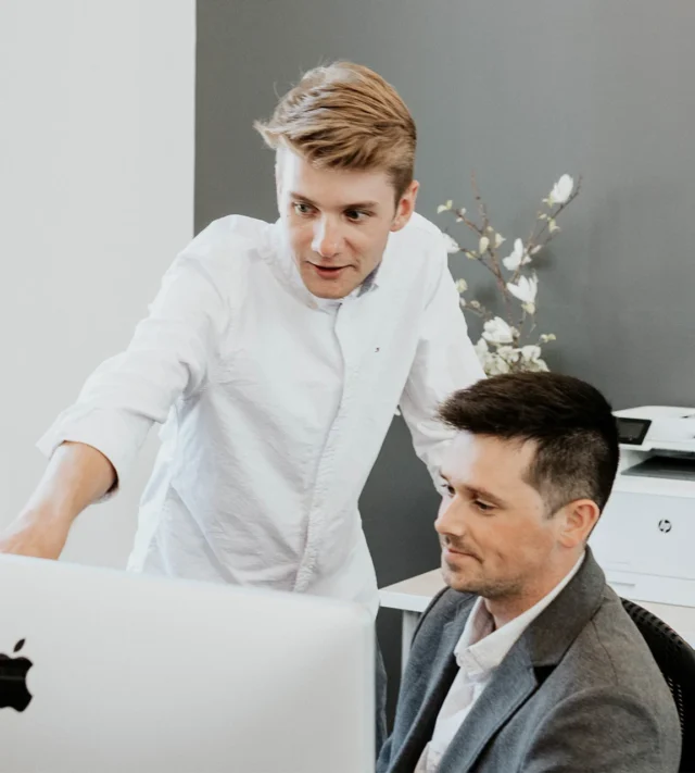 Two individuals in an office setting, one standing and pointing at an Apple computer screen, the other seated and looking at the screen.