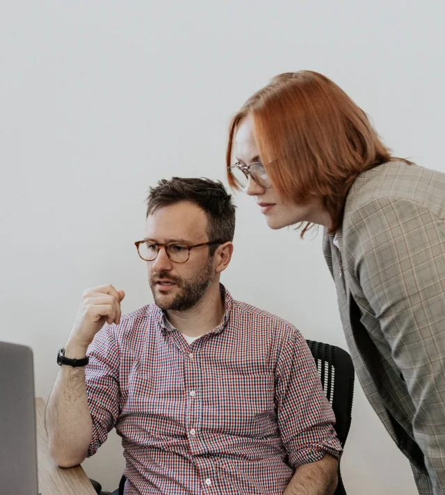 Two people in a meeting room look intently at a computer screen. The seated person gestures while the other stands and leans in, both wearing glasses.