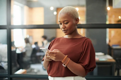 Person with short hair using a smartphone in an office setting, standing by a glass partition with people working in the background.