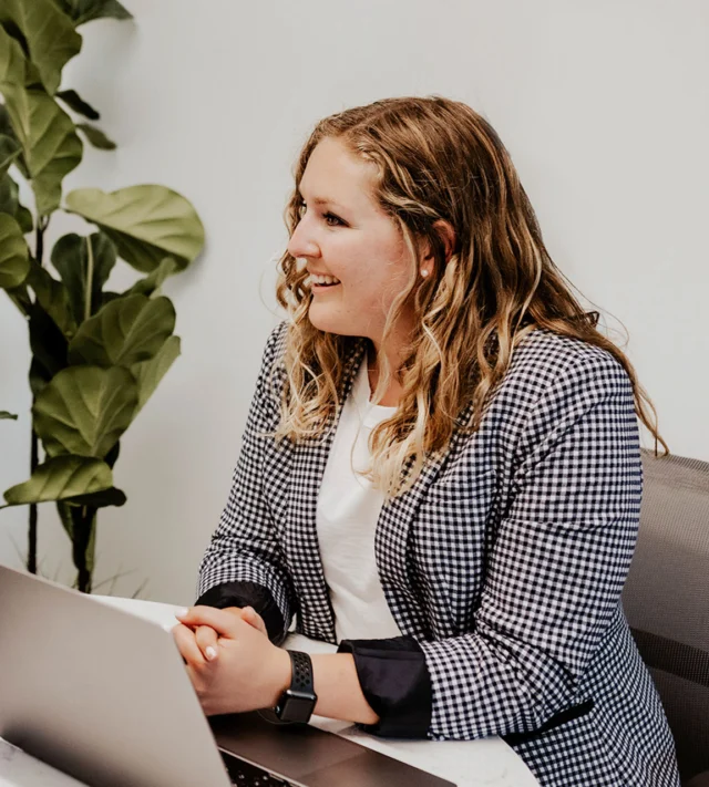 Person with long hair, in a checked blazer, sits at a desk with a laptop, smiling. There's a plant in the background against a white wall.