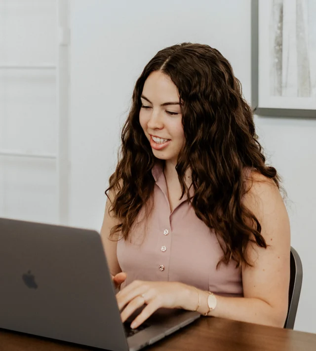 Person with long curly hair using a laptop at a table, wearing a sleeveless top and smiling. There is a framed picture on the wall in the background.