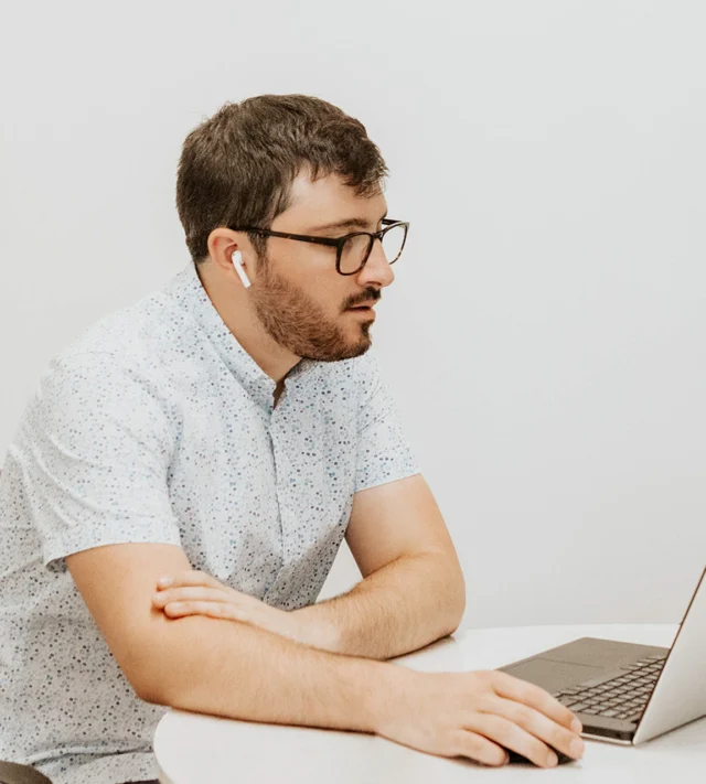 Man with glasses wearing earphones, sitting at a table, using a laptop.