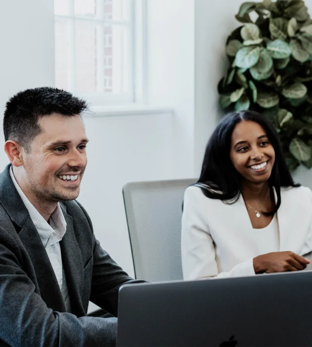 Two people in business attire smiling, seated at a table with a laptop, in an office setting with a large window and a plant in the background.