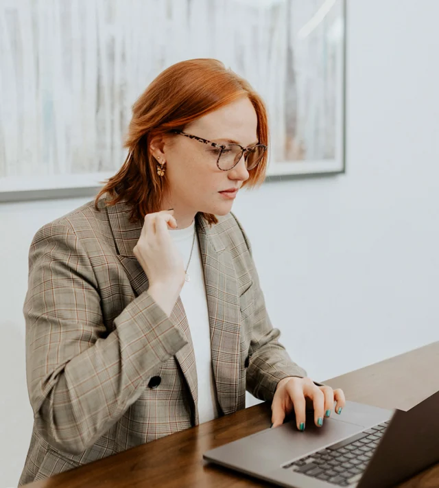 Person with red hair in glasses and plaid blazer, sitting at a desk, typing on a laptop.