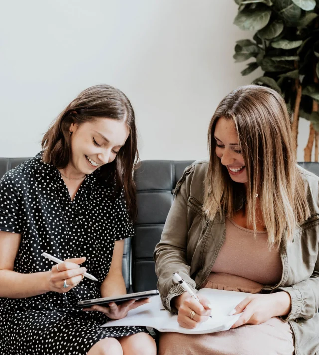 Two women sitting on a couch, smiling and writing notes. One holds a tablet with a stylus, while the other writes on a notepad. There's a plant in the background.