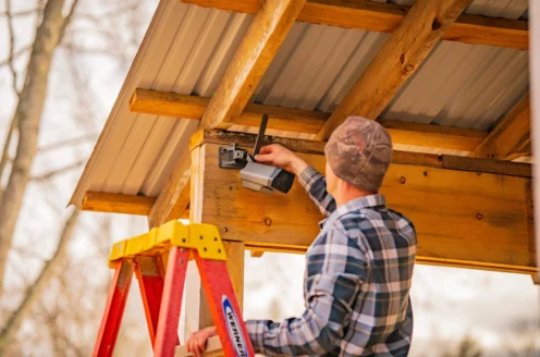 Person on a ladder installing a security camera under a wooden and metal roof.
