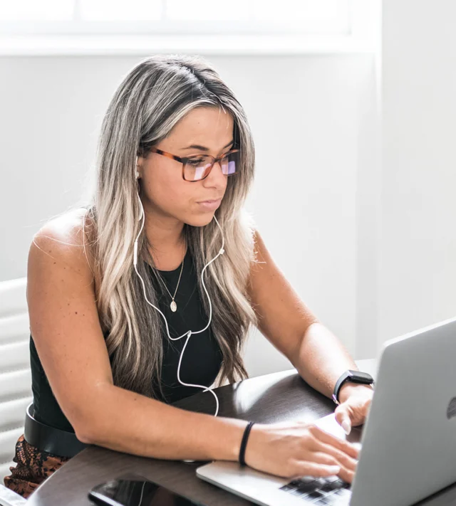 A woman with long hair and glasses is sitting at a table using a laptop. She is wearing earphones and a black tank top.