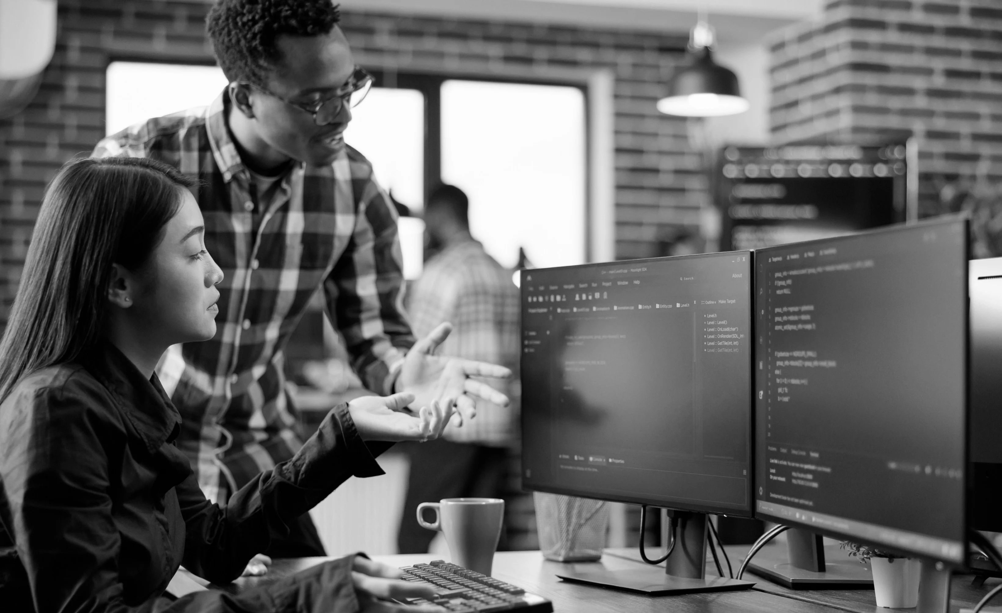 Two people work together at a computer desk. The woman types while the man gestures towards the screen. There are dual monitors displaying code. A coffee mug is on the table.