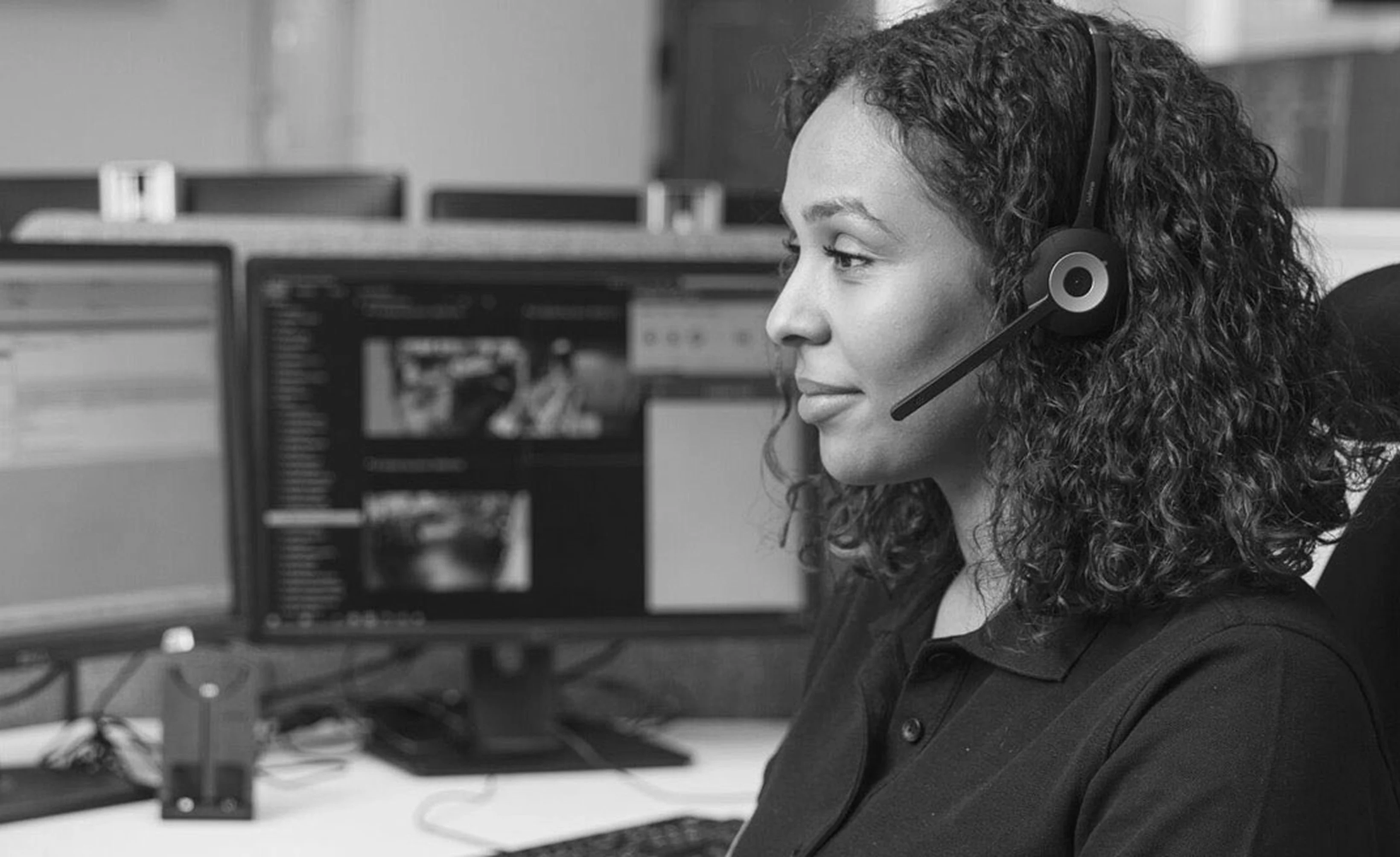 A woman with curly hair wearing a headset sits at a desk with multiple computer screens in the background.